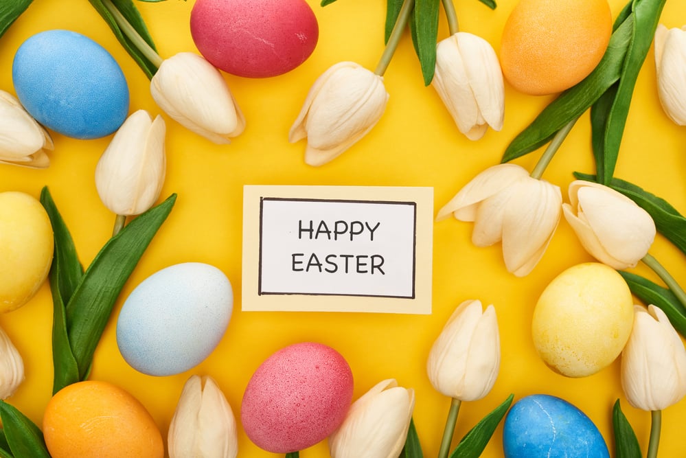 Happy Easter written on paper on yellow background surrounded by tulips and eggs