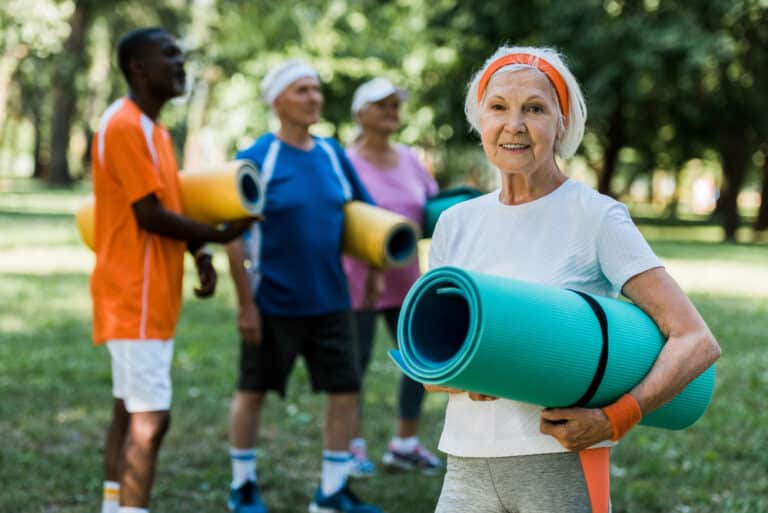 Senior woman holding rolled up yoga mat and smiling, friends behind her