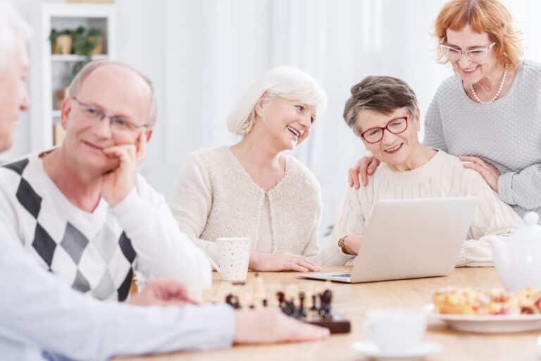 Three senior women smiling and looking at laptop, senior man chatting with friend